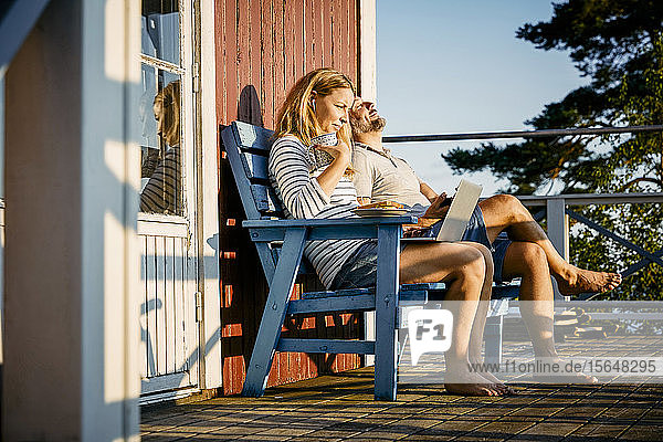 Mittlere erwachsene Frau  die während des Frühstücks einen Laptop benutzt und mit dem Mann auf der Veranda sitzt