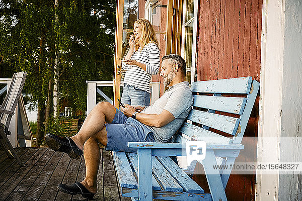 Lächelnder Mann sitzt auf Bank  während die Frau auf der Veranda mit dem Handy telefoniert
