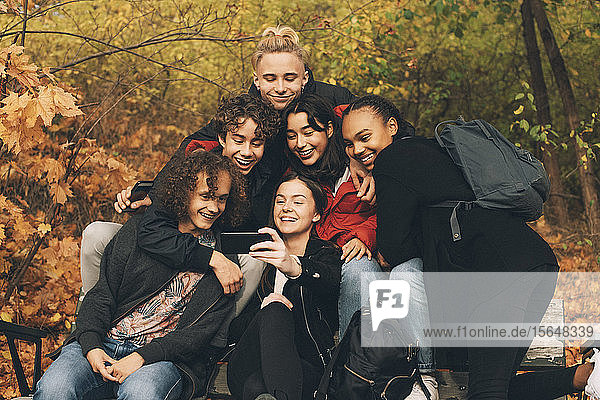 Teenage girl taking selfie with friends while sitting on bench against trees during autumn