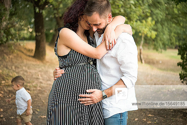 Man hugging and touching belly of pregnant wife  son in background in park