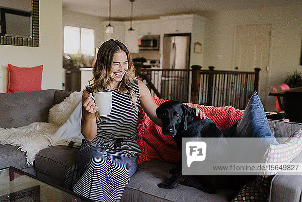 Frau genießt Tasse Heißgetränk  streichelt Hund auf dem Sofa