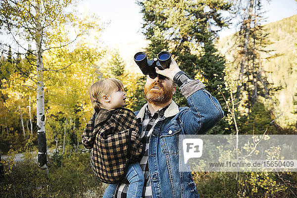 Mann hält seine Tochter im Arm  während er ein Fernglas im Wald benutzt