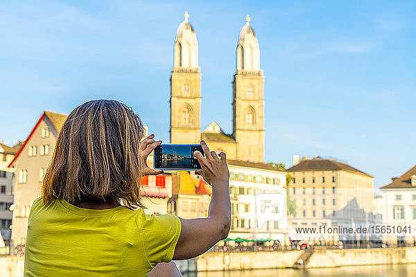 Rückansicht einer Frau  die das Grossmünster und den Fluss Limmat mit einem Smartphone fotografiert  Zürich  Schweiz