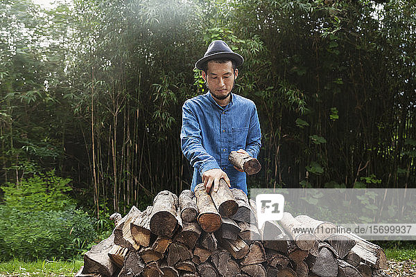 Japanese man wearing hat standing outdoors  stacking logs of firewood.