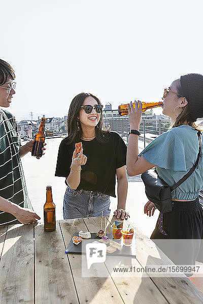 Ein junger Japaner und zwei Frauen stehen auf einem Dach in einer städtischen Umgebung und trinken Bier.