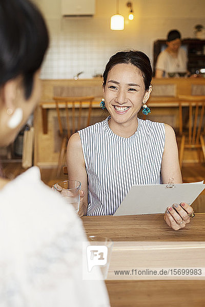 Lächelnde Japanerin  die an einem Tisch in einem vegetarischen Café sitzt und die Speisekarte hält.