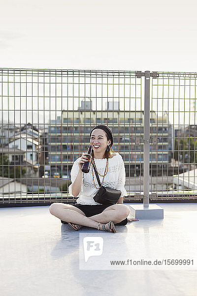 Lächelnde junge Japanerin  die in einer städtischen Umgebung auf einem Dach sitzt und Bier trinkt.