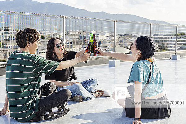 Ein junger Japaner und zwei Frauen sitzen auf einem Dach in einer städtischen Umgebung und trinken Bier.