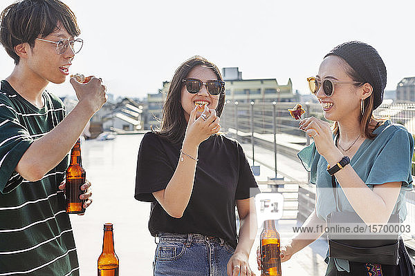 Ein junger Japaner und zwei Frauen stehen auf einem Dach in einer städtischen Umgebung und trinken Bier.