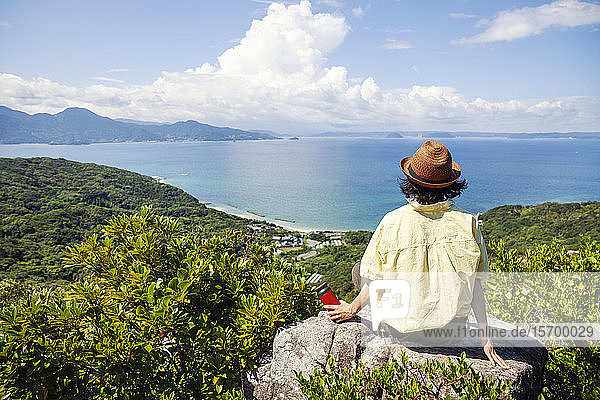 Japanische Frau mit Hut  die auf einem Felsen auf einer Klippe sitzt  im Hintergrund das Meer.