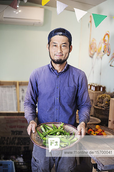 Lächelnder Japaner mit Mütze steht im Hofladen und hält eine Schüssel mit frischer Okra in der Hand.