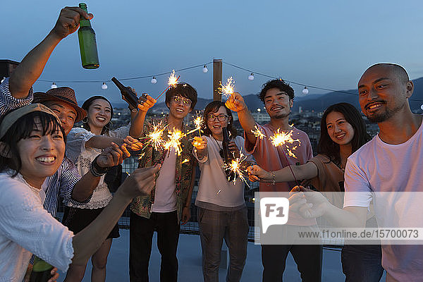 Group of young Japanese men and women with sparklers on a rooftop in an urban setting.