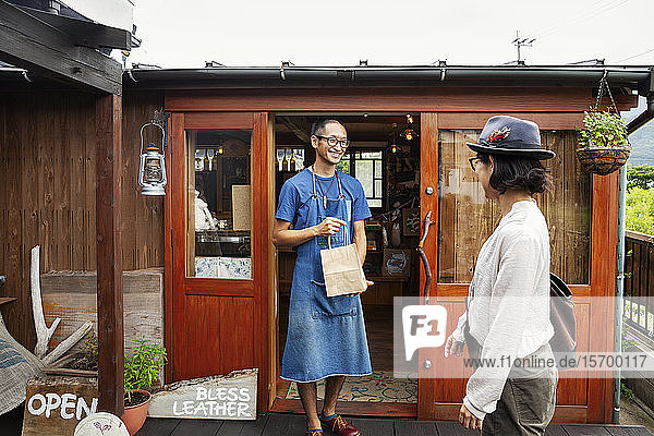 Japanese woman and man wearing blue apron and glasses standing outside a leather shop  holding shopping bag.