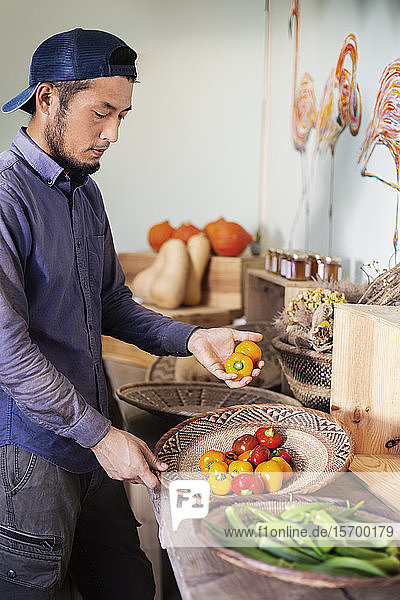 Japanese man wearing cap standing in farm shop  holding bowl with fresh vegetables.