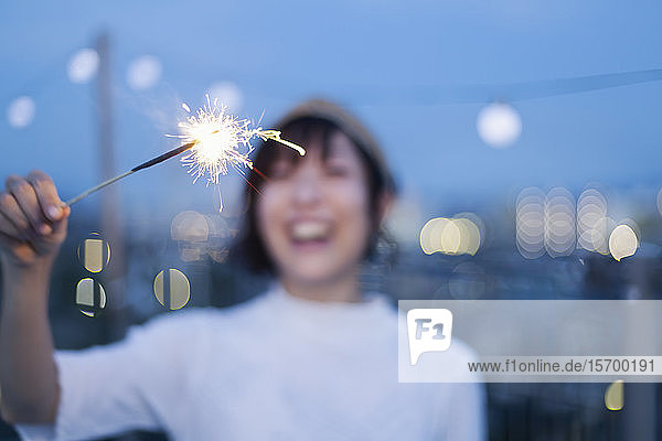Lächelnde junge Japanerin mit einer Wunderkerze in der Hand auf einem Dach in einer städtischen Umgebung.