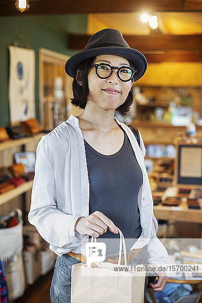 Japanerin mit Hut und Brille steht in einem Ledergeschäft  hält Einkaufstasche und lächelt in die Kamera.