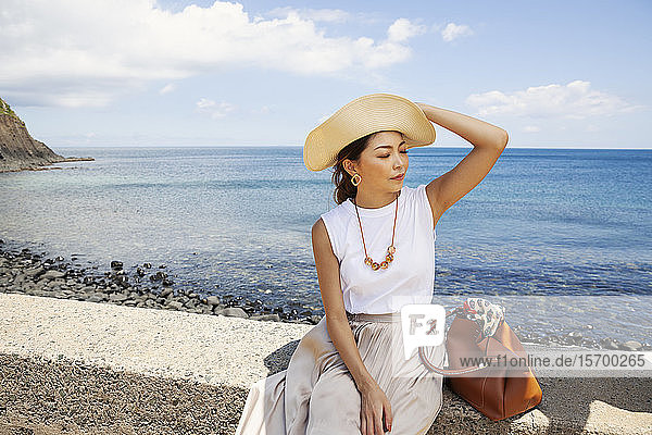 Japanese woman wearing hat sitting on a wall  ocean in the background.