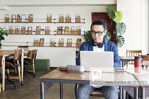 Männlicher japanischer Berufstätiger  der an einem Tisch in einem Raum für Zusammenarbeit sitzt und einen Laptop-Computer benutzt.