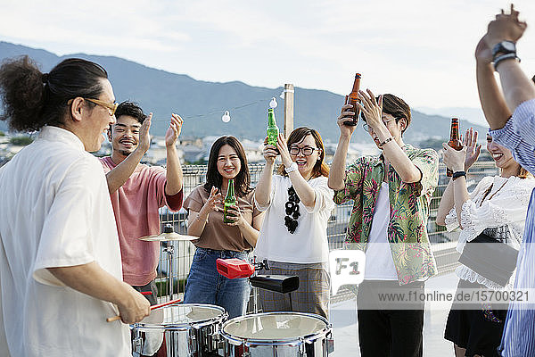Lächelnde Gruppe junger japanischer Männer und Frauen  die auf einem Dach in einer städtischen Umgebung stehen und Bier trinken.