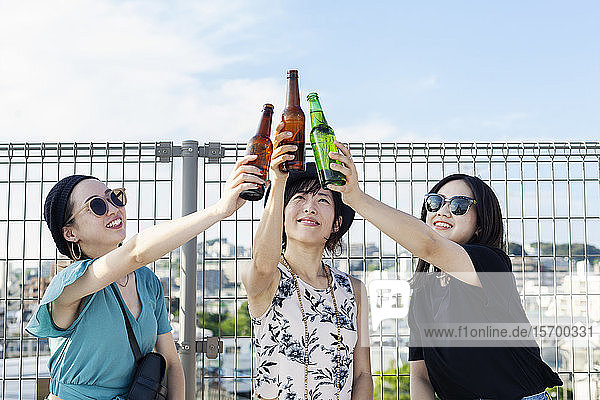 Drei junge Japanerinnen sitzen auf einem Dach in einer städtischen Umgebung und trinken Bier.