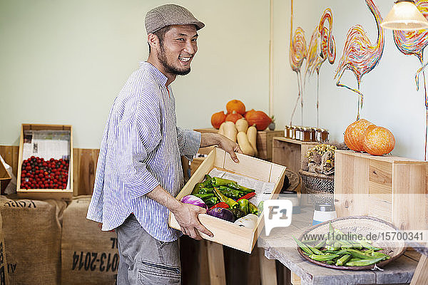 Lächelnder japanischer Mann mit Mütze steht im Hofladen und hält eine Kiste mit frischem Gemüse.