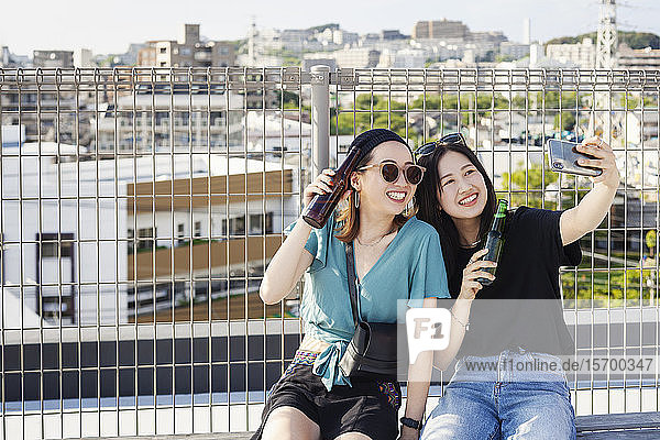 Two young Japanese women sitting on a rooftop in an urban setting  taking selfie with mobile phone.