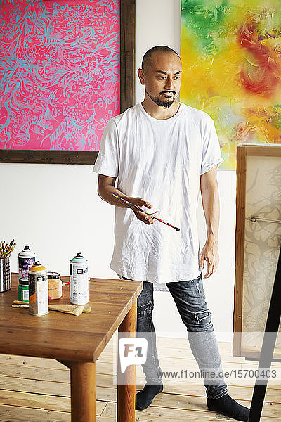 Japanischer Mann steht in der Kunstgalerie  hält Pinsel und betrachtet Kunstwerke auf der Staffelei.