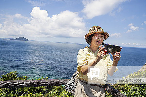 Japanese woman wearing hat standing on a cliff  taking selfie with mobile phone  ocean in the background.