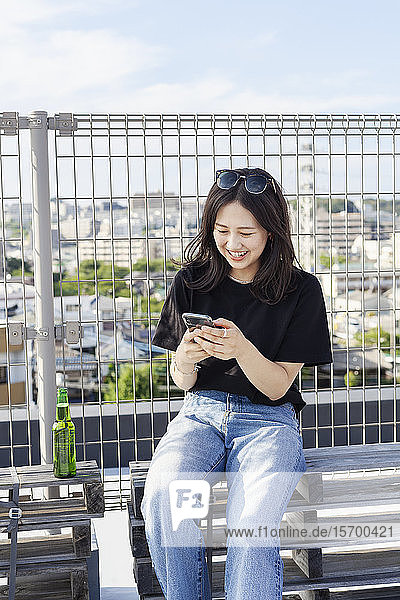 Junge Japanerin  die in einer städtischen Umgebung auf einem Dach sitzt und ein Mobiltelefon benutzt.