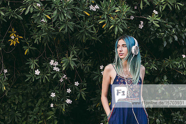 Junge Frau mit blauen Haaren hört Musik mit Kopfhörern vor einem blühenden Baum