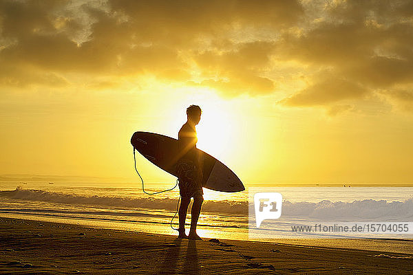 Silhouettierter männlicher Surfer mit Surfbrett am ruhigen Strand bei Sonnenuntergang