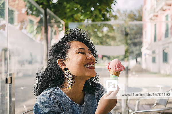 Unbekümmerte,  aufgeregte junge Frau,  die auf der Terrasse eines sonnigen Cafés eine rosa Eiswaffel genießt