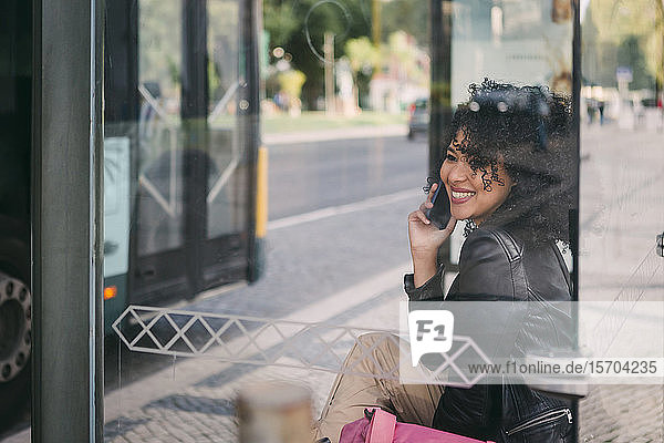 Glückliche junge Frau  die an der Bushaltestelle mit ihrem Smartphone telefoniert