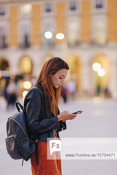 Young woman with backpack using smart phone on urban sidewalk