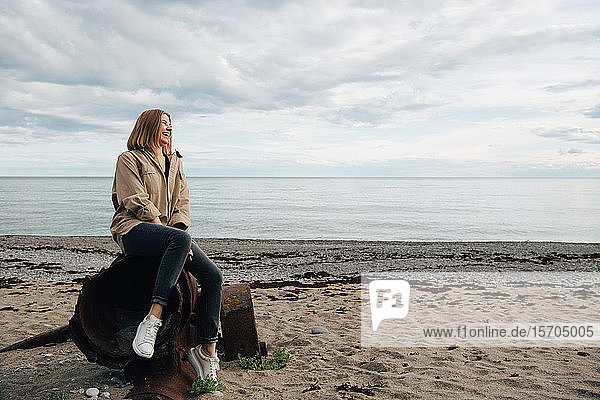 Cheerful young woman sitting on weathered metal at beach against sky