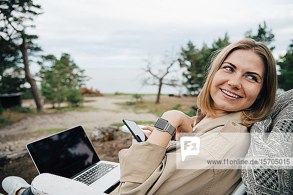 Lächelnde junge Frau mit Technologien schaut weg  während sie in den Ferien auf einem Stuhl sitzt
