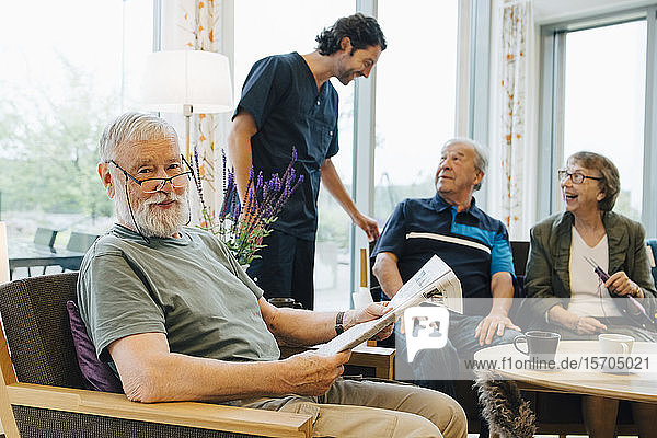 Portrait of retired senior man sitting with newspaper on armchair by friends and caretaker at elderly nursing home