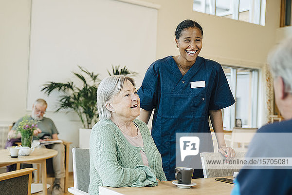 Cheerful young female nurse standing by senior woman while looking at man in retirement home