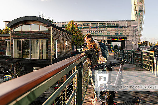 Couple with electric push scooters enjoying on bridge in city against sky
