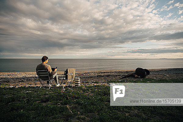 Mann in voller Länge mit Laptop und Mobiltelefon  während er am Strand vor bewölktem Himmel sitzt