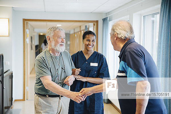 Älterer Mann Arm in Arm mit Krankenschwester  während er einem Freund in einer Gasse im Pflegeheim die Hand schüttelt
