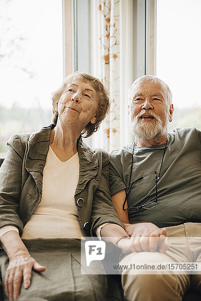 Älteres Ehepaar hält sich an den Händen  während es am Fenster eines Pflegeheims sitzt