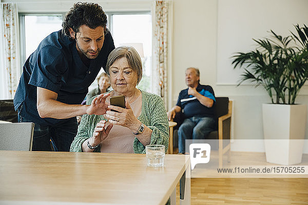 Zuversichtlicher männlicher Pfleger unterstützt ältere Frau bei der Nutzung eines Smartphones im Pflegeheim