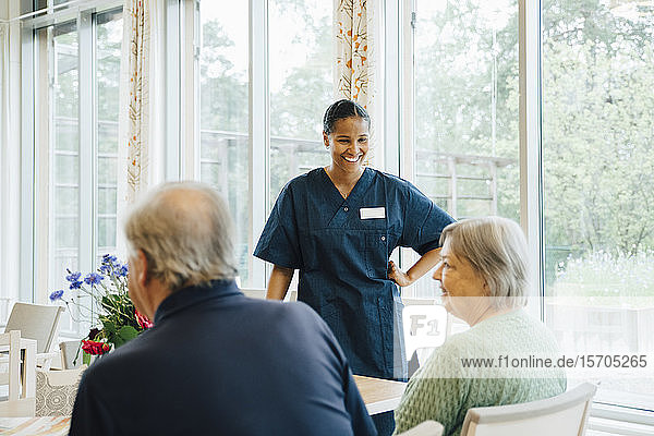Lächelnde junge Krankenschwester sieht ältere Frau an  die von einem Mann am Esstisch im Altenpflegeheim sitzt