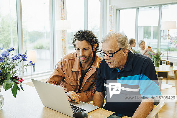 Mann assistiert Großvater mit Laptop am Esstisch im Altersheim