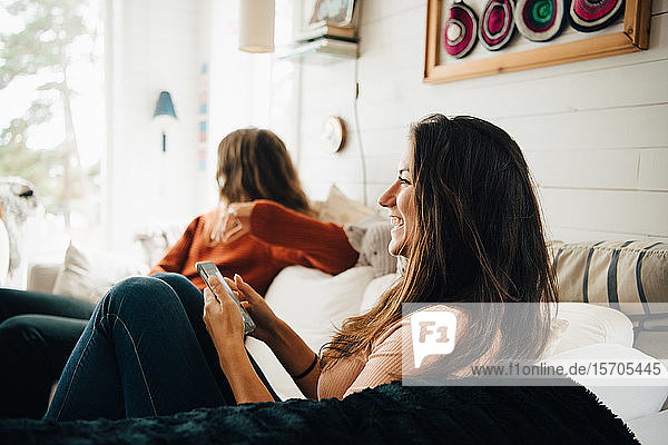 Smiling woman with smart phone looking away while sitting with friend on sofa at home