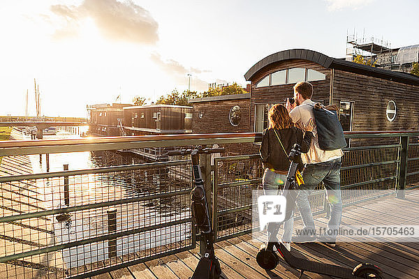 Paar mit Elektroschubroller fotografiert Fluss, während es auf einer Brücke in der Stadt steht