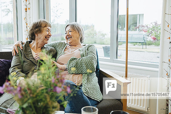 Lächelnde ältere Frauen sitzen mit umarmtem Arm und schauen sich auf dem Sofa gegen das Fenster im Altersheim an