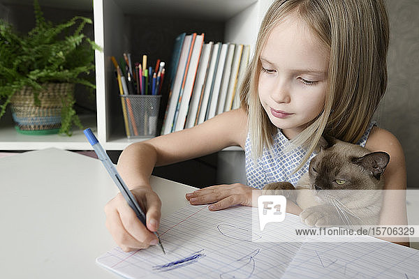 Girl with a cat sitting at table at home doing homework