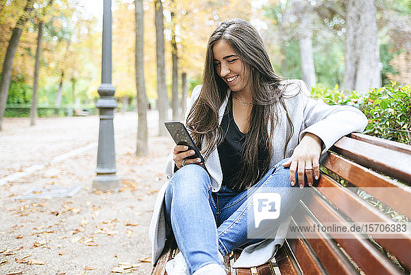 Lächelnde Frau sitzt auf einer Parkbank und benutzt ein Smartphone